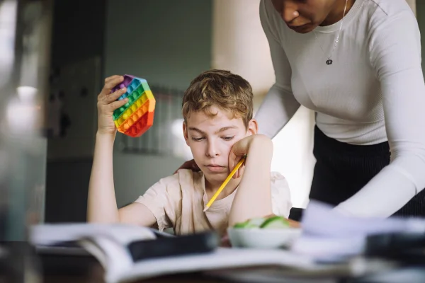 Foto mostra uma criança branca fazendo o dever de casa enquanto segura um fidget pop-it colorido que representa do TDAH - Metrópoles