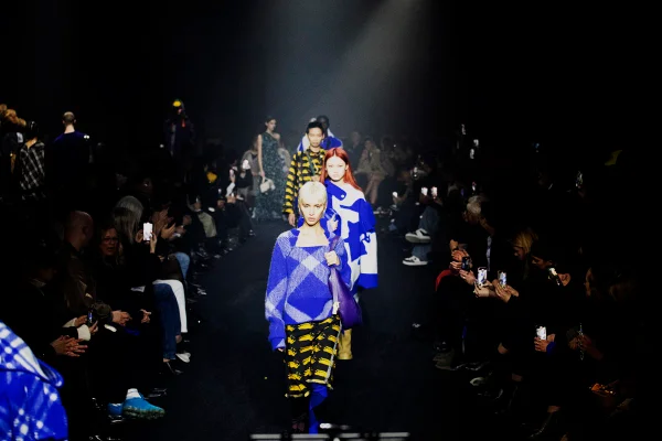 Em desfile, fila de modelos em meio à plateia. A modelo da frente usa look estampado em xadrez com blusa e suéter em azul e branco, além de saia em amarelo e preto - Metrópoles