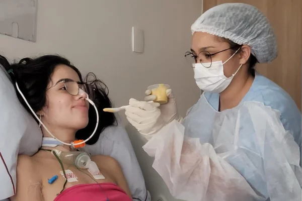 Foto colorida de jovem sobre uma cama de hospital, conectada a fios, recebendo de uma enfermeira comida na boca. -Metrópoles.