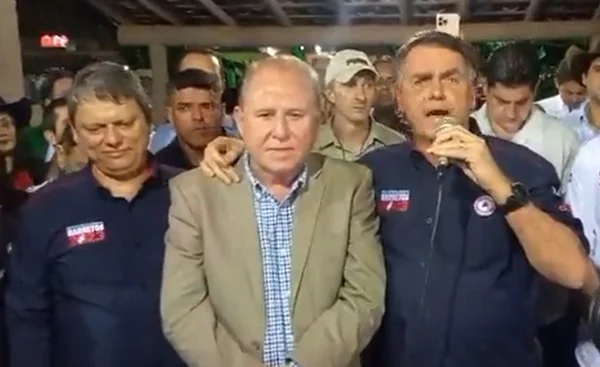 Foto colorida do governador Tarcísio de Freitas à esqueda de camisa azul escura com o ex-presidente Jair Bolsonaro à direita de camisa escura e falando ao microfone - metrópoles