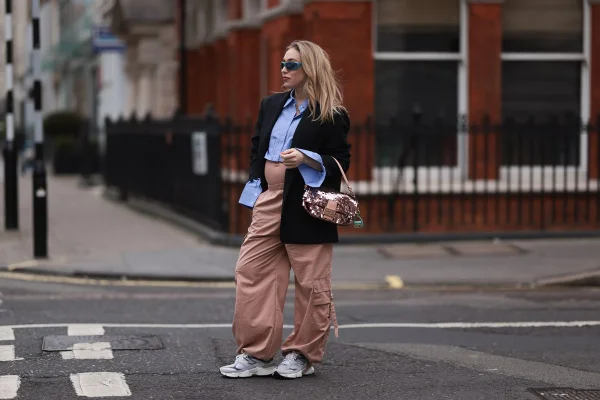No street style, mulher loira grávida usa camisa social azul com blazer preto, além de calça paraquedas em tom terroso - Metrópoles