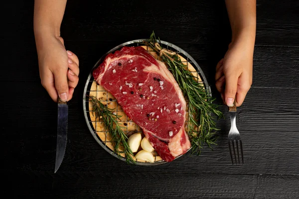 Foto colorida de um prato com um pedaço de carne, pouco temperada e crua, com folhagens ao redor -Metrópoles.