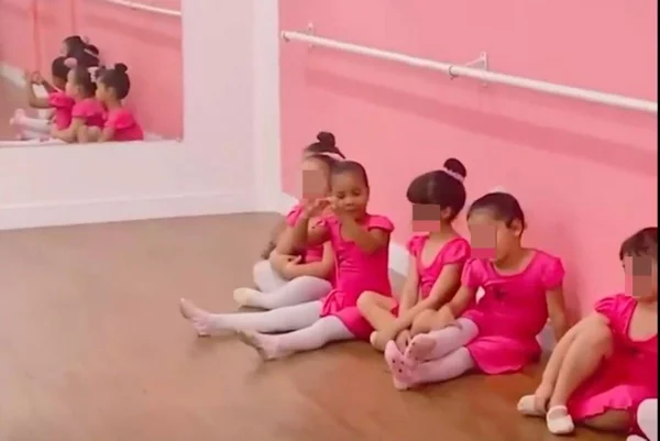Imagem colorida de meninas sentadas em sala de balé, com parede rosa ao fundo, crianças vestem vestido rosa e meia-calça branca. Uma menina faz símbolo de coração com as mãos - Metrópoles