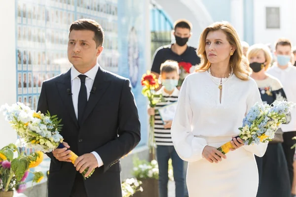 A primeira-dama da Ucrânia, Olena Zelenska, com roupas brancas ao lado do marido, o presidente Zelensky, de terno. Eles carregam flores - Metrópoles