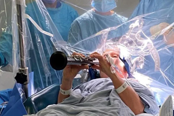 Foto colorida de mulher deitada numa mesa de cirurgia, em volta panos azuis e um instrumento de sopro na maõ - Metrópoles.