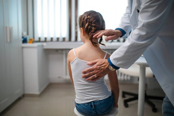 Imagem colorida de adolescente com escoliose sentada em maca médica sendo analisada - Metrópoles