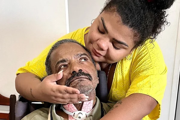 Arlindo Cruz e Flora Cruz. O cantor está sentado em uma cadeira e utliza um aparato no pescoço. Flora está de camisa amarela e com o cabelo preso - Metrópoles