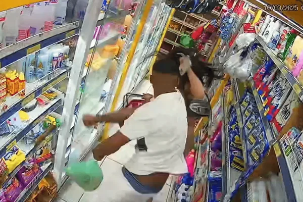 Imagem colorida de uma câmera de segurança mostra um homem de camiseta branca agredindo com socos uma mulher vestida de bege no corredor de um mercado - Metrópoles