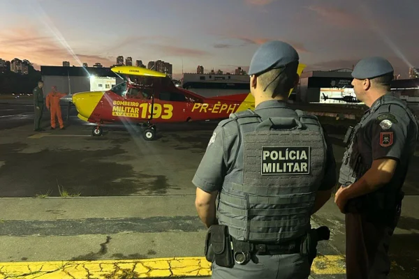 Imagem colorida mostra dois policiais militares de costas olhando para aeronave ao fundo - Metrópoles