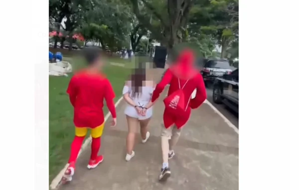 Imagem colorida de dois homens vestidos com fantasias vermelhas de Chapolin Colorado escoltando mulher branca com shorts e camisa brancos