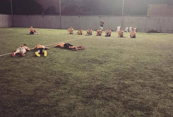 foto colorida de uma campo de futebol com três homens deitados no chão, oito sentados e um em pé