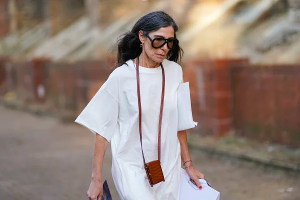 Mulher branca, de meia idade, com cabelos escuros lisos e cumpridos, e alguns fios brancos. Ela usa óculos escuros; uma pequena bolsa marrom pendurada no pescoço para celular; e um vestido branco.