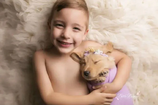 FOTO DE MENINO DEITADO EM TAPETE DE PELINHOS SEGURANDO CACHORRO EMBRULHADO EM PANO ROXO