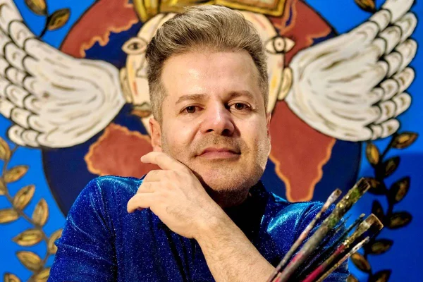 foto colorida de homem branco posando com pincéis e quadro ao fundo - metrópoles