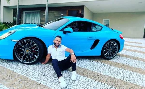 Foto colorida de Felippe Ribeir, da empresa Avanzza, sentado no chão, de camisa branca, calça preta em frente a um carro de luxo azul - metrópoles