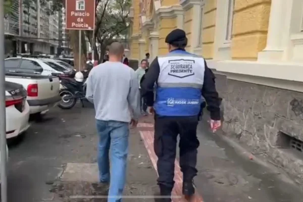 Foto colorida mostra um policia da Segurança Presente conduzindo ex-pm preso por suposta tentativa de matar o próprio irmão no Rio