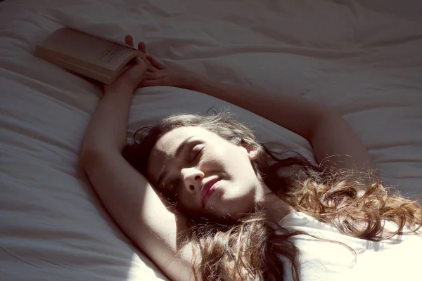 Foto colorida de uma mulher deitada na cama com a luz da janela batendo em seu rosto com o livro na mão - Metrópoles