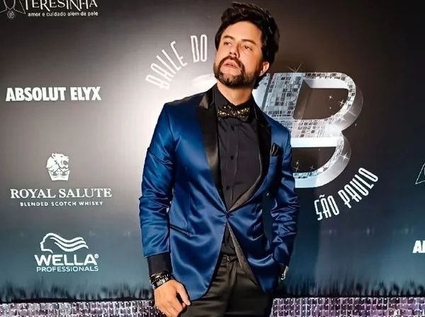 Foto colorida mostra o ator Rafael Suco, um homem branco, com cabelo preto para cima, barba, vestindo um terno azul brilhante e uma camiseta preta por baixo - Metrópoles