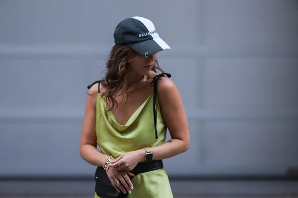 Mulher branca com cabelo comprido castanho e ondulado. Ela usa um smartwatch da marca apple, um vestido verde de alcinha e um boné preto e branco.