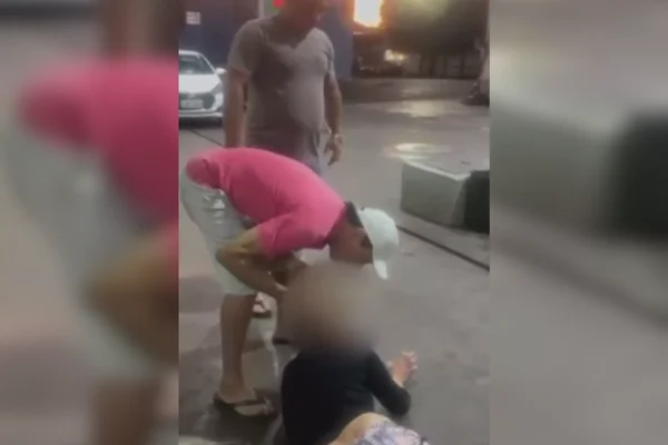 Imagem colorida de um homem de boné branco agredindo uma mulher, sem reação, jogada no chão de um posto de gasolina - Metrópoles