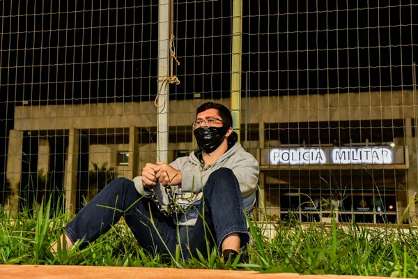 Imagem de um homem sentado no chão, vestindo calça jeans azul, casaco cinza e tênis pretos. Ele está acorrentado pelos braços junto a uma grade cinza, sentado de costas para a grade, com óculos e máscara no rosto. A grade é das dependências do QG da PMDF