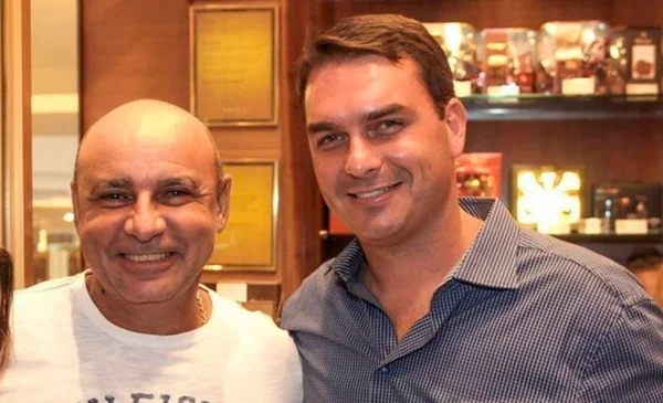 Na imagem colorida, dois homens estão posicionados no centro. Eles sorriem para a camera