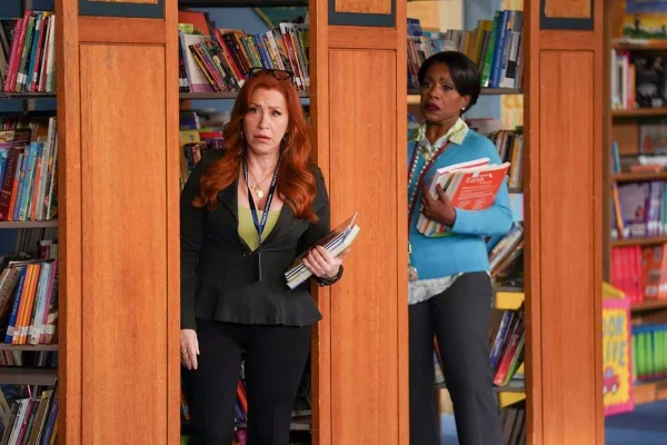 duas mulheres seguram livros em corredores de biblioteca e olham chocadas para algo fora da imagem - metrópoles