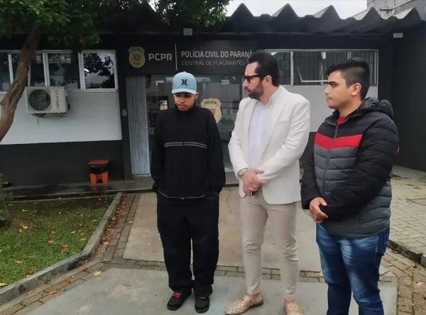 Imagem colorida mostra frentista vítima de racismo, vestido de moletom preto e boné azul, ao lado de seu advogado, de terno claro, e outro homem, de casaco, em frente a delegacia em Curitiba - Metrópoles