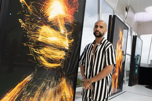 Gabriel Wickbold está com camisa e calça listradas em preto e branco. Ele está perto de uma de suas obras - Metrópoles