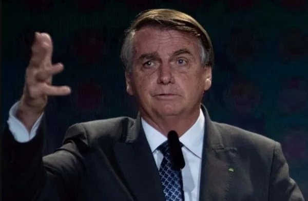 Na imagem colorida. Um homem está posicionado no centro. Ele usa terno e gravata, tem cabelos curtos e grisalhos e olha atentamente para a frente