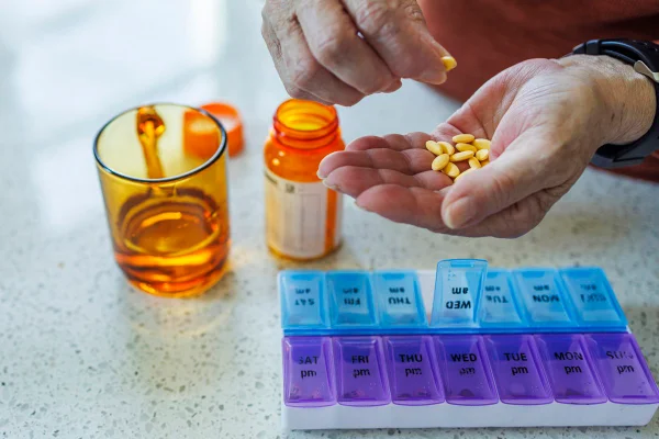 Foto colorida de mão segurando cápsulas de medicamentos e copo de água - Metrópoles