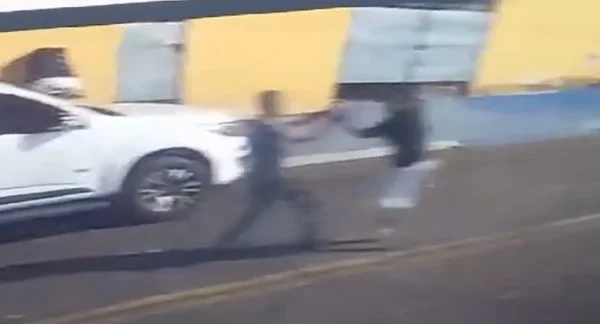 Imagem colorida mostra dois homens brigando na frente de um carro branco. O idoso de calça comprida acabou levando um tiro do jovem de bermuda - Metrópoles