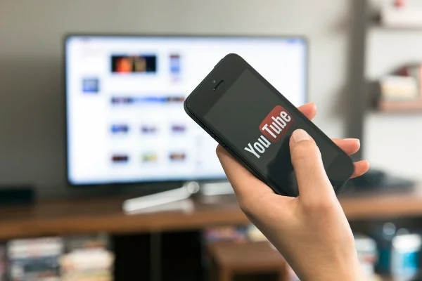 Pessoa segurando um celular com logo do YouTube, aparecendo televisão ao fundo - Metrópoles