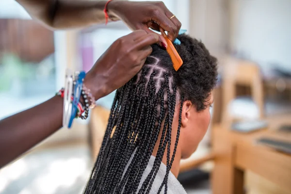 Uma mulher negra de meia-idade trança o cabelo de uma mulher mestiça em um ambiente doméstico, demonstrando a confecção de um penteado tradicional. Metrópoles
