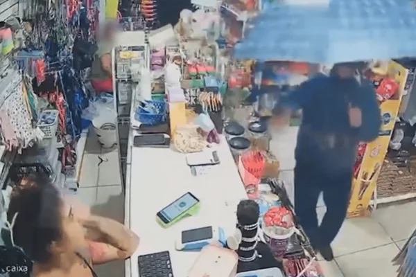 Imagem colorida de um homem encapuzado segurando um guarda-chuva azul e um revólver dentro de um pet shop. O homem aparece mirando para uma atendente de um petshop - Metrópoles