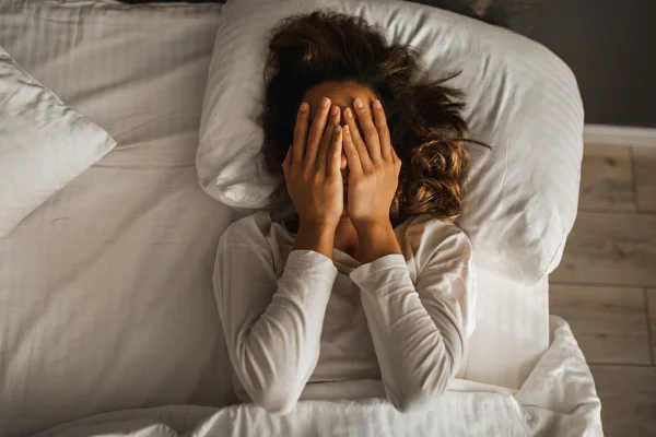 Foto colorida de mulher deitada em cama e com as mãos no rosto - Metrópoles
