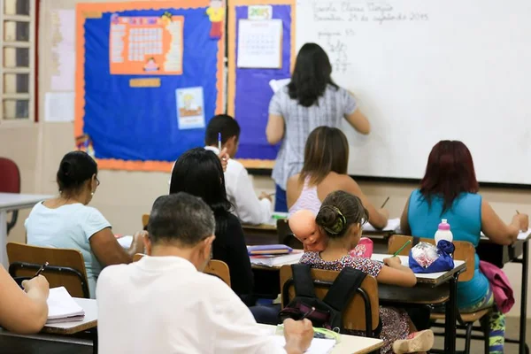 Estudantes sentados em carteiras virados para o quadro e mulher escrevendo em quadro branco