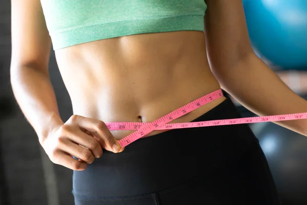 Foto colorida de mulher com fita métrica na região abdominal. Ela usa um top cropped verde e uma calça preta - Metrópoles