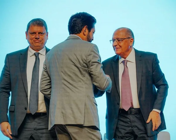 Imagem colorida mostra Geraldo Alckmin e Ricardo Nunes, ambos de terno e gravata, se cumprimentando, na frente de um fundo azul. A cena tem uma terceira pessoa, Tarcísio de Freitas, também de terno