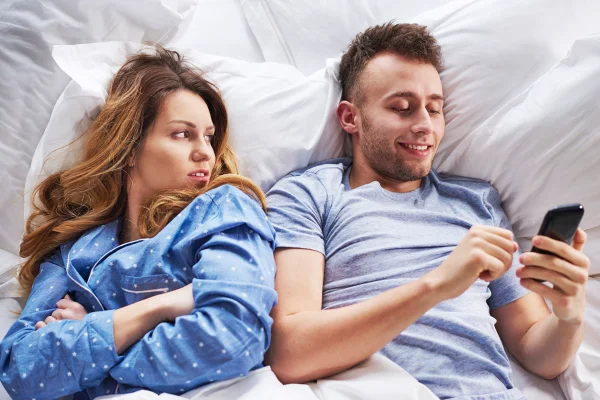 Foto colorida de mulher com cara de indignação ao ver namorado mexer no celular na cama