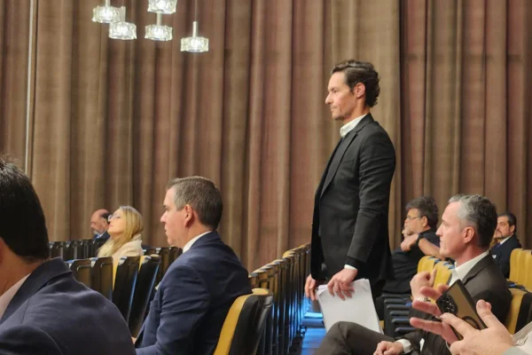 imagem colorida mostra homem em pé com documentos na mão olhando para frente. ele está em uma sala com cortinas marrons e cadeiras amarelas. ao seu redor, outras pessoas também olham para frente