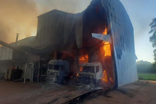 Imagem colorida de caminhões pegando fogo dentro de galpão em chamas. Metrópoles