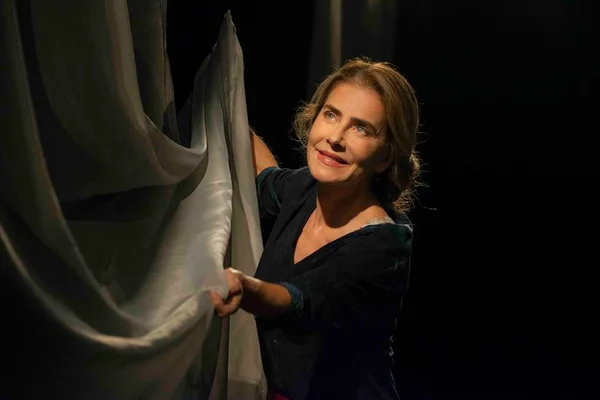 Foto colorida de Maitê Proença com um vestido preto segurando uma cortina de seda cinza - Metrópoles