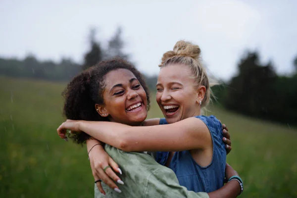 Imagem colorida de duas mulheres se abraçando enquanto riem em campo verde aberto - Metrópoles