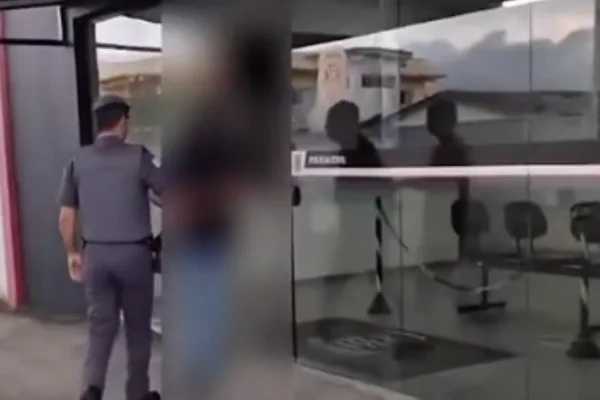 Imagem colorida de policial militar conduzindo preso algemado à delegacia. Metrópoles