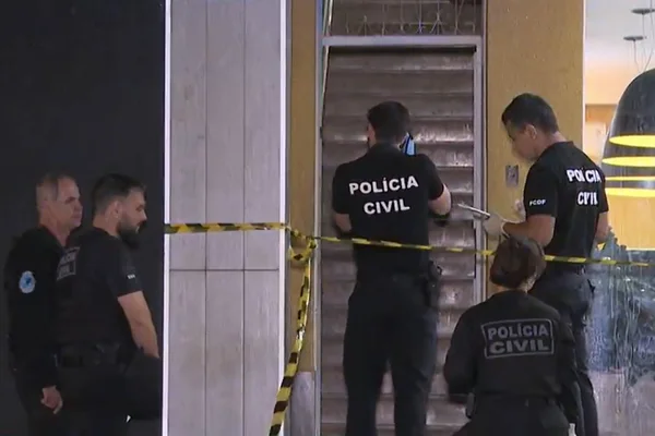 Imagem colorida mostra equipes da Polícia Civil, com uniformes pretos, em frente a entrada de prédio, em área isolada