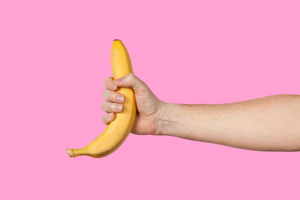 foto colorida de uma mão masculina segurando uma banana com casca em um fundo cor-de-rosa - Metrópoles