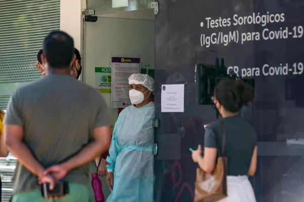 mulher com materiais de proteção - touca, roupa de proteção e máscara, conversando com dois homens que estão em uma longa fila