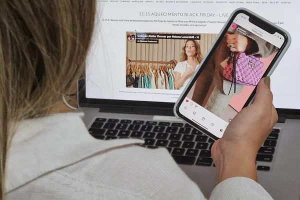 Mulher branca e jovem mexendo no celular e no computador. Ela olha sites de compra de roupas e acessórios