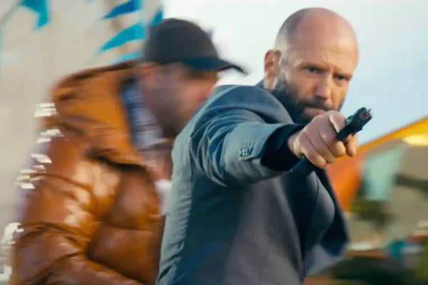 Foto de um homem branco e careca de terno cinza atirando com uma pistola - Metrópoles